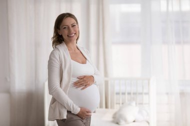 Müstakbel anne portresi konsepti. Modern çocuk odasında poz veren 30 'lu yaşlarda hamile güzel bir kadın hamile. Hamileliğin sonlarına doğru karnına dokun. Kameraya bakıp gülümse.
