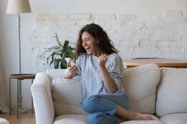 Neşeli mutlu Latin güzel bir kadın cep telefonu tutuyor, gülüyor, bağırıyor, kazanan jestler yapıyor. Heyecanlı cep telefonu kullanıcısı rahat koltukta oturup başarıyı kutluyor.