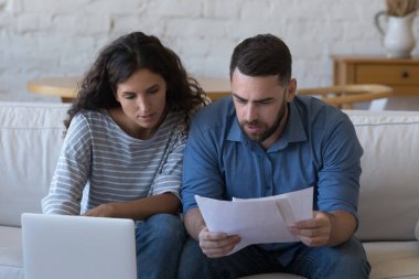 Odaklanmış evli çift dizüstü bilgisayarda kağıt okuyor, gayrimenkul satın alma sözleşmesini gözden geçiriyor, ipotek, sigorta sözleşmesi, banka tebligatı, birlikte evrak işi yapıyor.
