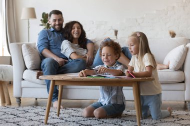 Küçük kız kardeşine kalem çizmeyi öğreten, gülümseyen, gülen, karalayan mutlu bir anaokulu çocuğu. Neşeli ebeveynler evde küçük çocuklarla vakit geçiriyorlar.