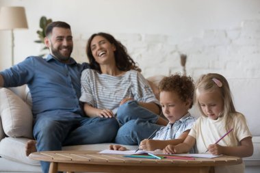 Neşeli genç ebeveynler rahat evlerinde kanepede dinleniyor, kağıt albümde renkli karalamalar çizen iki çocuğu izliyorlar, renkli kalemler kullanıyorlar, oturma odasında oynuyorlar, aile eğlencelerinin tadını çıkarıyorlar.