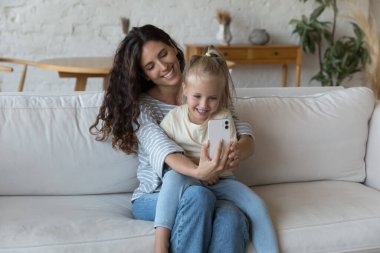 Mutlu blogcu anne kucağında neşeli, tapılası küçük bir kızı kucağında tutuyor, akıllı telefonda selfie çekiyor, video konuşmasında konuşuyor, yumuşak koltukta, cep telefonu ekranında dinleniyor.
