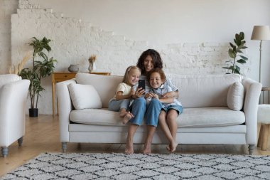 Neşeli, güzel bir anne cep telefonuyla koltukta dinleniyor ve iki küçük mutlu çocuk, şık, rahat bir oturma odasında selfie çekiyor, görüntülü konuşuyor, aile tatilinin tadını çıkarıyor, internet iletişiminin tadını çıkarıyor.