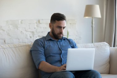 Evde çalışan, rahat koltukta oturan, dizüstü bilgisayarı kucağında tutan, daktilo eden, ekrana bakan, kablosuz internet iletişimi için aygıt kullanan odaklı dizüstü bilgisayar kullanıcısı.