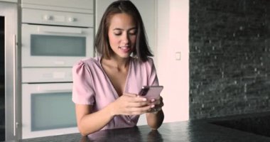 Pozitif odaklı genç Asyalı kadın cep telefonuyla sohbet ediyor, gülümsüyor, modern ev mutfağında cihazlara yazı yazıyor, kurye, uygulama, iletişim için modern kablosuz internet teknolojisi kullanıyor.