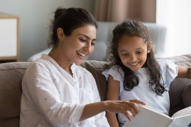 Gülümseyen Hintli kadın küçük kızına kitap okurken, anaokulu 6 'lı yaşlarındaki sevimli kız kanepeye oturur ve annelerin masal anlatarak evde vakit geçirmesinin keyfini çıkarır. Çocuk gelişimi, hobi, iyi yaşam alışkanlığı