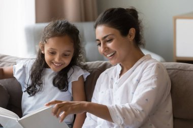 Gülümseyen Hintli kadın küçük kızına kitap okurken, anaokulu 6 'lı yaşlarındaki sevimli kız kanepeye oturur ve annelerin masal anlatarak evde vakit geçirmesinin keyfini çıkarır. Çocuk gelişimi, hobi, iyi yaşam alışkanlığı