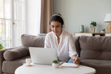 Hintli bir kadın elinde kalem tutarak not alıyor, ödev yazıyor, egzersiz yapıyor, yabancı dil çalışıyor, evde bilgisayar kullanarak e-öğreniyor. Uzaklık çalışması için video arama uygulaması kullanımı