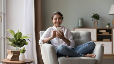 Genç gülümseyen Hintli kadın, elinde modern cep telefonuyla koltukta oturmuş kameraya bakıyor. Mutlu bir kadın iletişim kurmaktan hoşlanır, mesajları ve mesajları paylaşır, akıllı telefon aygıtını kullanır.