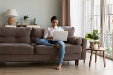 Ciddi Hintli kadın kucağına kablosuz bilgisayar koydu müşteriye e-posta attı oturma odasındaki rahat kanepeye oturdu. Freelancer evde çalışıyor, tele-iş odaklanmış görünüyor. Modern teknoloji kullanımı