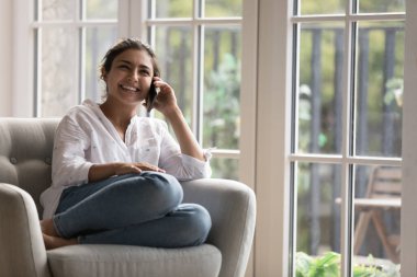 Gülümseyen Hintli kadın evinde koltuğunda oturmuş akıllı telefonuyla hoş sohbetler yapıyor, konuşuyor, haberleri paylaşıyor, modern teknoloji, bağlantı ve telefon kullanarak iletişimden keyif alıyor.