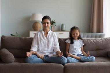 Hintli kadın ve kız evdeki kanepede Lotus pozisyonunda oturmuş meditasyon yapıyorlar, sakin gözlerle gözlerini kapatıyorlar birlikte oturma odasında yoga çalışmalarının keyfini çıkarıyorlar. Ebeveynler çocuklarına iyi yaşam alışkanlığı öğretir