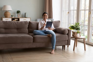 Genç Hintli kadın kanepede dinleniyor, evde zaman geçiriyor, tablet tableti kullanıyor, kameraya bakıyor, modern cihaz kullanımının keyfini çıkarıyor, uzaktan sipariş veriyor, internette eğleniyor, sosyal medya ve yeni uygulama kullanıcısı arıyor.