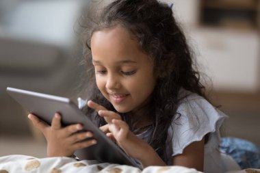 Şirin Hintli anaokulu öğrencisi kız internette dijital tablet cihazıyla yatarak vakit geçiriyor, kapat. Gelişim ya da eğlence için yeni uygulama kullanımı, modern kablosuz teknolojiye bağımlı çocuk, kötü alışkanlık