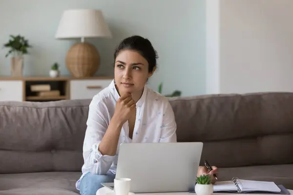 stock image Pensive Indian woman holds pen thinks over ideas staring into distance while sitting on sofa with laptop and personal diary looking thoughtful, deep in thoughts, search solutions and creative decision