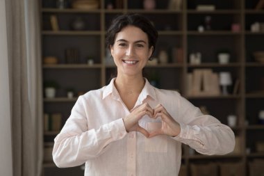 Mutlu güzel Latin kız göğsünde el ele tutuşmuş, kamera karşısında parmak kalp şeklinde. Genç blogcu kadın aşkı, ilgiyi, desteği, minnettarlığı ifade ediyor, jest yapıyor, kameraya bakıyor, gülümsüyor.