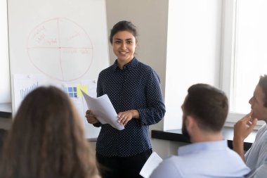 Kendine güvenen Hintli iş kadını projeyi meslektaşlarına sunuyor, şirket toplantısında çalışan ekiple konuşuyor, elinde kağıt belgeler tutuyor. Kadın akıl hocası eğitim grubu