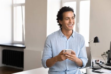 Mutlu düşünceli genç iş adamı elinde cep telefonu tutuyor, internet iletişimi için internet uygulamasını kullanıyor, sohbet ediyor, çalışma masasında duruyor, başka yere bakıyor, düşünüyor, hayal ediyor, gülümsüyor.