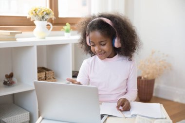 Pembe kulaklıklı mutlu siyah nesil ilkokul çocuğu online derse katılıyor, evde bilgisayarla ilgili sanal dersler alıyor, öğrenim programlarını izliyor, ders çalışmaktan, kazanmaktan, internetteki eğitimden keyif alıyor.