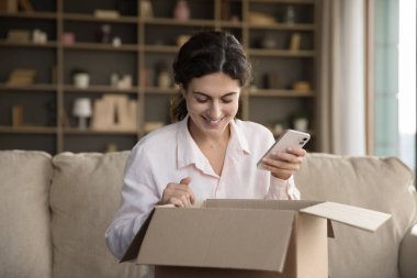 Neşeli müşteri kız internet mağazasından satın alıyor, cep telefonundaki uygulamayı kullanıyor, dükkana geri bildirim veriyor. Müşteri kadın paket paketini evdeki kanepede açıyor, cep telefonu kullanıyor. Teslimat konsepti