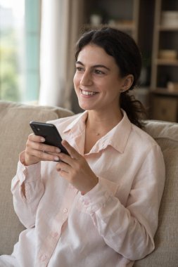 Mutlu cep telefonu kullanıcısı kız, evde kanepede oturuyor, dişlek bir gülümsemeyle başka tarafa bakıyor, mesaj yazıyor, daktilo ediyor, mesaj yazıyor, akıllı telefon kullanıyor. Dikey çekim