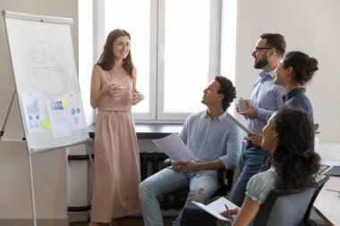 Mutlu bağlı şirket koçu istatistik verilerini grafik üzerinde sunuyor. İş adamları pazarlama raporunu tartışıyor, analitik grafikler üzerinde fikir alışverişinde bulunuyor.