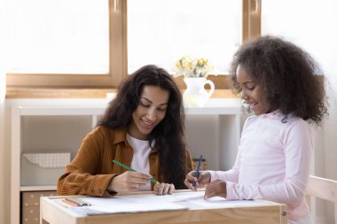 Neşeli kreş öğretmeni kağıt albümde kalemlerle resim çizen küçük mutlu okul kızını izliyor. Afrikalı anne, tatlı kızına ödevlerinde yardım ediyor. Çocukluk, çocuk eğitimi konsepti