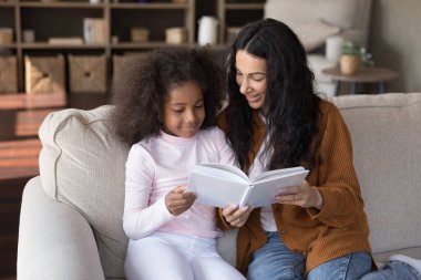 Mutlu bir anne çocuğuna okumayı öğretiyor, açık bir kitap tutuyor, kanepede oturuyor, okul ödevi için kıza yardım ediyor, yardım ediyor, eğitim veriyor, edebiyata ilgi duyuyor.