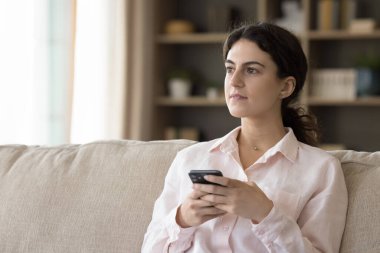 Milenyum kızı akıllı telefon tutuyor, rahat bir ev koltuğunda oturuyor, pencereye bakıyor, internetten sohbet ediyor, mesajlaşıyor, mesajlaşıyor, sanal satın alma, alışveriş yapıyor.