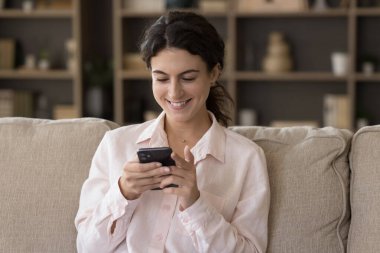Milenyum müşterisi genç bir kız akıllı telefon kullanıyor, oturma odasındaki koltukta dinleniyor, internetten alışveriş yapıyor, cep telefonundan sanal ekommerce uygulaması alıyor, mesaj yolluyor.