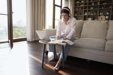 Bin yıllık İspanyol öğrenci pembe kablosuz kulaklıklı kız öğrenim notları yazıyor, okul ödevi yapıyor, ödev yazıyor, kompozisyon yazıyor, açık kitaplarda dizüstü bilgisayarda Webinar izliyor.