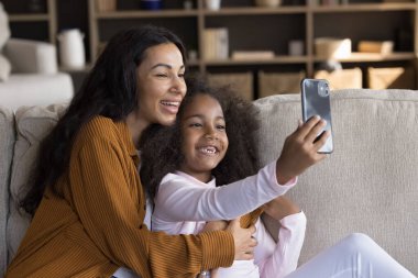 Mutlu Siyahi anne ve küçük kızı cep telefonuyla selfie çekiyorlar, koltukta dinleniyorlar, eğleniyorlar, blog için komik bir mesaj kaydediyorlar, video konuşmaları yapıyorlar, internetten medya izliyorlar.