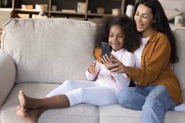 Mutlu genç anne ve çocuk akıllı telefondan video görüşmesi yapıyor, kanepede birlikte dinleniyorlar, internetten cep telefonu alışverişi yapıyorlar. Z jenerasyonundaki kıza hücre ekranında çevrimiçi öğrenme uygulaması gösteriliyor