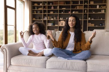 Mutlu, barışçıl Afrikalı anne ve kız evdeki kanepede gözleri kapalı meditasyon yapıyor, el zen parmaklarını tutuyor, sakin, sakin bir gülümseme takınıyor. Anne kızına yoga ve zihinsel egzersiz yapmayı öğretiyor.