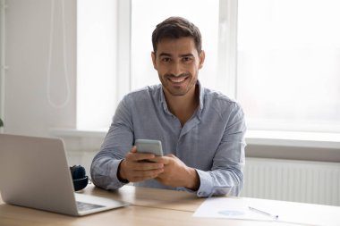 Akıllı telefon kullanan mutlu dijital alet kullanıcısı, iş yerindeki portrede laptop kullanıyor. Genç yakışıklı iş adamı cep telefonu tutuyor, bilgisayarın başında oturuyor, kameraya bakıyor.