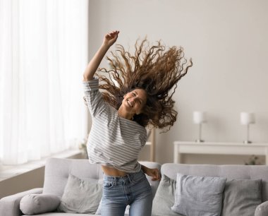 Uzun kıvırcık saçlarıyla dans eden, müzikten hoşlanan, aktif boş vakitleri olan, gözleri kapalı, modern evdeki dişlek gülümsemesi olan gamsız, zayıf, esmer, genç bir kadın.,