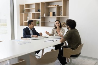 Yaşlı İspanyol kadın profesyonel finans danışmanı genç Hintli erkek girişimcilere brifing konusunda yardım ediyor. Çeşitli şirket çalışanları beyin fırtınasında sinerji ile işbirliği yapıyor.