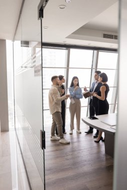 Ciddi yöneticilerden oluşan çok ırklı bir ekip modern ofis toplantılarında çalışma alanlarını tartışıyor, cam duvarın arkasındaki işbirliği odasında duruyor, konuşuyor, beyin fırtınası yapıyor. Dikey çekim