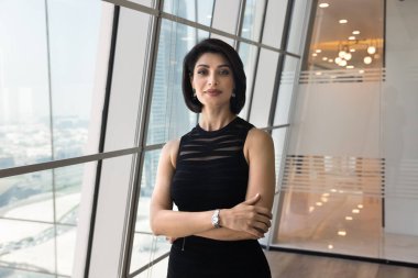 Başarılı esmer iş kadını. Elleri bağlı kameraya bakıyor. Geniş penceredeki modern kentsel ofis çalışma alanında profesyonel portre için poz veriyor.