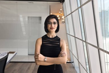 Kendine güvenen, gözlüklü, ellerini ofisin içinde çapraz durarak poz veren, güven jesti yapan, kameraya bakan, kendine güvenen genç ve profesyonel bir kadın. Başarılı kadın lider portresi