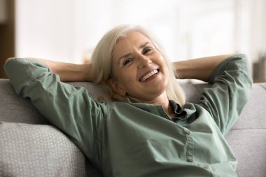 Kaygısız, olgun bir kadın ellerini başının arkasına koyar evde dinlenir, gülümser, kameraya bakar huzur ve sessizlik anının tadını çıkartır. Yeniden düzenle, sağlıklı gündüz istirahatı, stresten uzak.