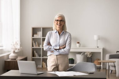 Şık giyinmiş, kendine güvenen, modern işyerinde kollarını kavuşturmuş, kameraya bakan, tek başına poz veren olgun bir iş kadını başarılı ve bağımsız görünüyor. Küçük işletme sahibi, şirket başkanı portresi.