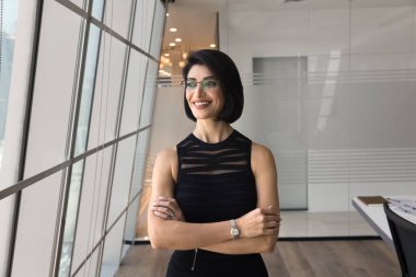 Kendine güvenen, gözlüklü genç bir iş kadını. Ofiste elleri bağlı duruyor. Pencereye mutlu düşüncelerle bakıyor, kariyer büyümesini planlıyor, şirketin başarısını düşünüyor.,