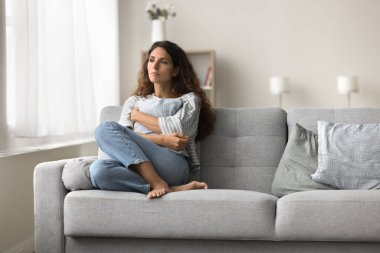 Hüzünlü, yalnız bir genç kadın modern iç mekanda koltukta oturuyor, kapalı poz veriyor, yastığı sıkıyor, depresyon geçiriyor, stres sonrası ilgisizlik, travma, kayıp.