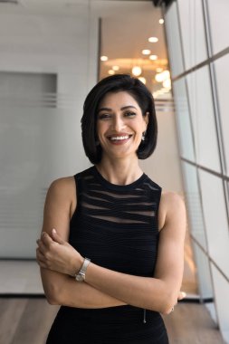 Neşeli, güzel, esmer, genç girişimci kadın, kollarını kendine güven jesti ile kavuşturuyor, kameraya bakıyor, gülümsüyor, gülüyor, iş başarısından keyif alıyor. Dikey portre