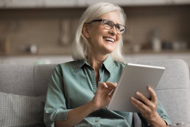 Modern tablet kullanan mutlu olgun bir kadın video görüşmeleri, sosyal medya, mesajlaşma uygulamaları, eğlenmek, çevrimiçi yayın hizmetleri veya e-kitaplar aracılığıyla eğlenmek için aile ve arkadaşlarla iletişimde kalmak için