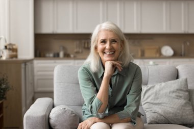 Orta yaşlı mutlu bir kadın modern bir dairede dinleniyor, gülümsüyor, koltukta tek başına oturan kameraya bakıyor. Tatmin olmuş, kaygısız, sağlıklı yaşlı bir kadının portresi emeklilikte boş ve sorunsuz bir hayatın tadını çıkarıyor.