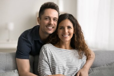 Pozitif yakışıklı koca, güzel karısına sarılıp koltukta arkadan oturuyor. Mutlu çift evde eğleniyor, dişlek tebessümle kameraya bakıyor, portre için poz veriyor.