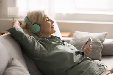 Yaşlı kadın rahat bir ifadeyle kanepede dinleniyor, kablosuz kulaklık takıyor, akıllı telefon tutuyor, favori müziğin tadını çıkarıyor, rahat görünüyor, rahatlık ve huzur dolu. Hobi, dinlen.