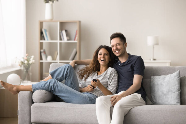 Joyful excited pretty couple spending leisure time at TV, using remote control, resting on comfortable home sofa, watching movie, sitcom show, comedy, online channel, hugging, laughing